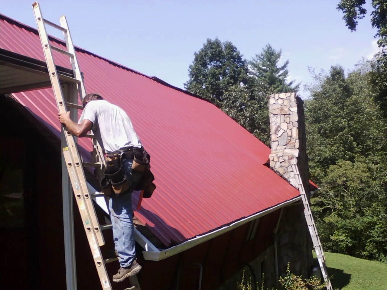 Roofer on a ladder beside a red metal roof and stone chimney — Soffit & Fascia Repair by Roofing Brothers in Carmel Valley